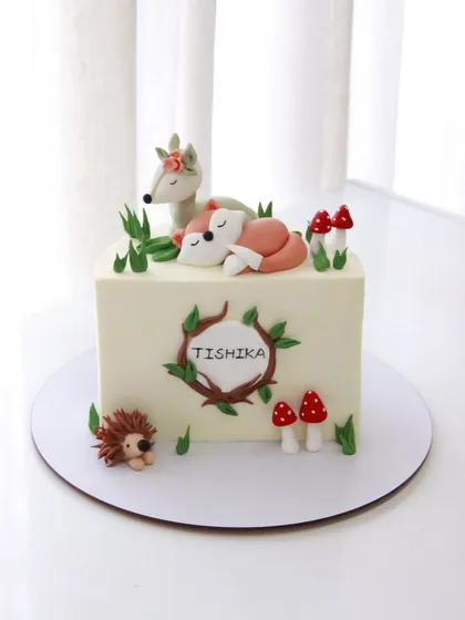 A whimsical woodland-themed half birthday cake for Tishika, featuring a sleeping fox, a deer, and a little hedgehog.