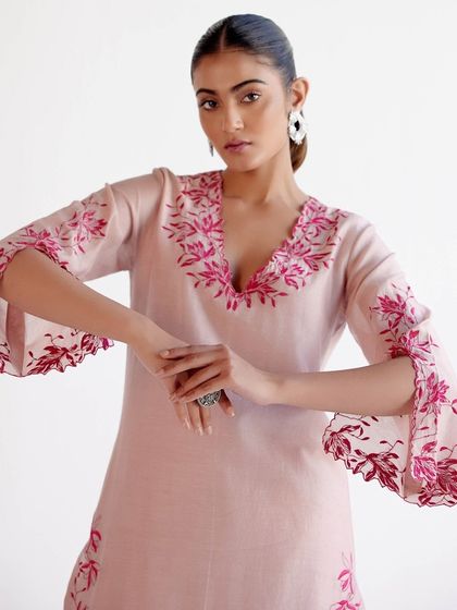 A model showcasing a light pink kurta with bell sleeves, adorned with delicate floral embroidery in shades of red.