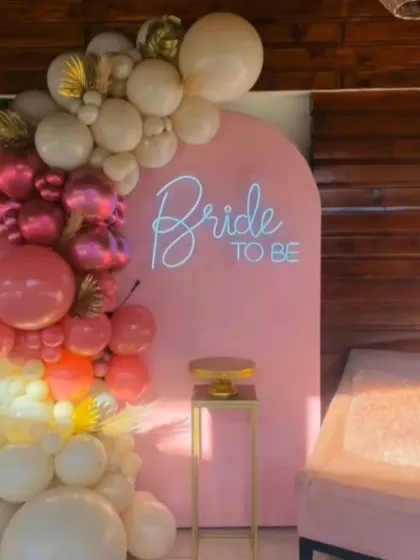 This modern "Bride to be" photo booth features a stylish pink arched panel with a custom neon sign. The look is completed with a cascading organic balloon garland in shades of pink, cream, and gold, perfect for creating memorable pictures.