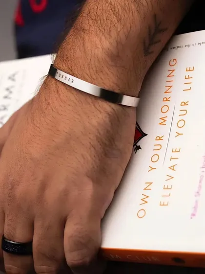 A man wearing a silver cuff bracelet engraved with the name 'Tushar'. This lifestyle shot shows how the minimalist design complements a casual, everyday look.