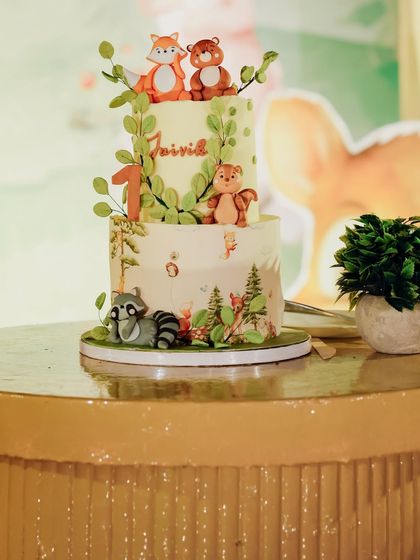 A lovely two-tiered woodland cake for Jaivik Viraj. It's adorned with cute fondant foxes and bears, surrounded by delicate green leaves, perfectly capturing the theme.