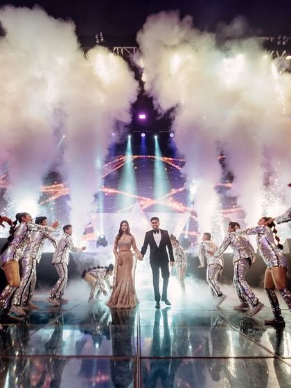 The walk-in moment. The couple emerges through a tunnel of our professional dancers and dramatic smoke effects, walking onto a reflective LED floor that adds another layer of visual grandeur to their sangeet entry.