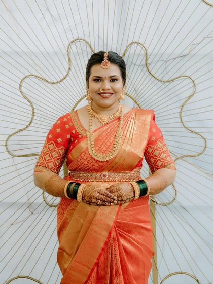 A happy bride in a beautiful orange Kanchivaram saree. The blouse features a geometric embroidery pattern on the sleeves, adding a modern touch.