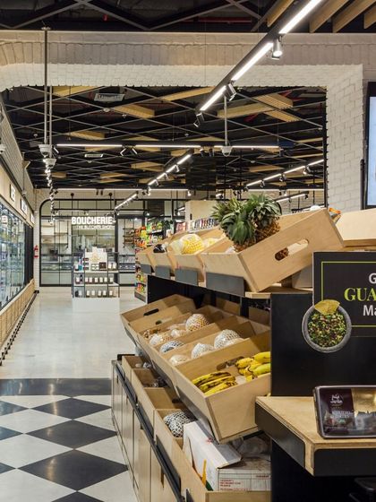 The produce section at Le Marche uses rustic wooden crates and focused lighting to highlight the freshness of the fruits and vegetables. In the background, a glimpse of the butchery section, framed by an arch, shows the store's organized, zone-based layout.
