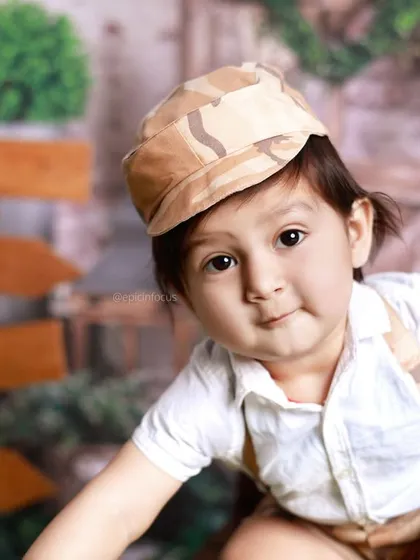 A charming close-up of a little boy in a camo cap, peeking at the camera. The outdoor setting allows for beautiful, naturally lit portraits.