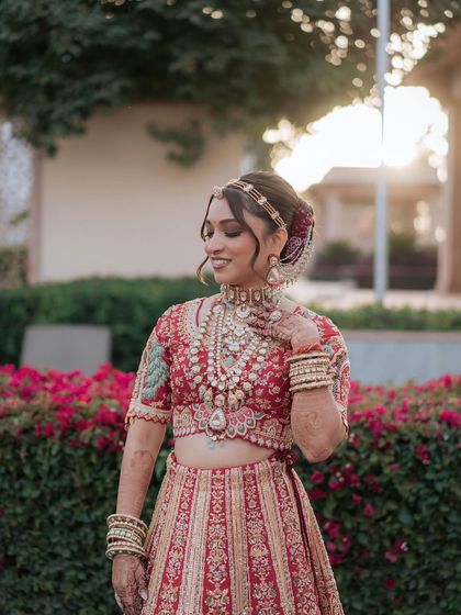 A portrait of the bride against a backdrop of flowers. The soft, romantic makeup and elegant hairstyle complement the intricate details of her red and gold lehenga.