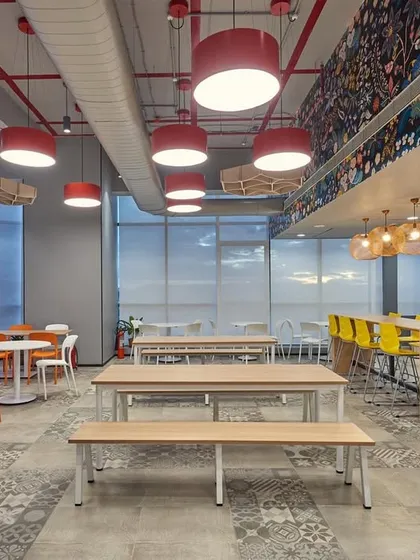 A large, bright cafeteria with a variety of seating options, including long communal tables, smaller round tables, and a high-top counter. The space is designed to accommodate a large number of people comfortably.