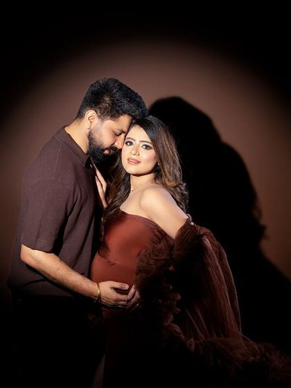 The deep, earthy tones of the gown and background create a warm, moody, and intimate portrait for this expecting couple.