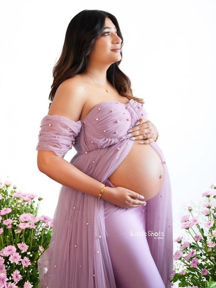 A beautiful portrait of an expecting mother in a lavender gown, set against a backdrop of pink flowers. Her off-the-shoulder gown elegantly drapes to reveal her baby bump.