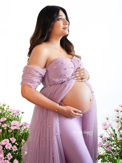 A beautiful portrait of an expecting mother in a lavender gown, set against a backdrop of pink flowers. Her off-the-shoulder gown elegantly drapes to reveal her baby bump.