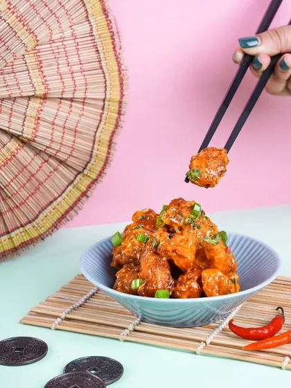 A hand using chopsticks to pick up a piece of crispy chili chicken. The playful, colorful background and props give this Indo-Chinese dish a fun and modern twist.