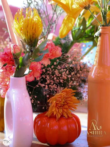 A close-up of a table centerpiece, with a mix of orange, yellow, and pink flowers in quirky, colorful vases.