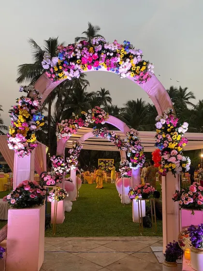 A vibrant, multi-colored floral archway for a daytime garden wedding. The mix of pinks, yellows, and blues creates a cheerful and festive entrance that is full of life.