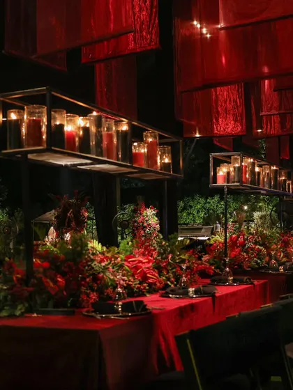 An intimate yet grand dining experience. Long tables are draped in red velvet, with overhead fabric installations and rows of candles creating a warm, romantic, and luxurious atmosphere for guests.