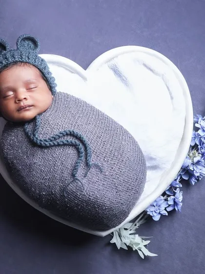 An overhead shot of a baby in a bear-eared hat, nestled in a heart-shaped bowl. The simple grey and blue color palette creates a calm and sweet image.