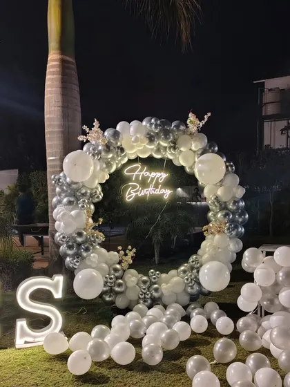 This outdoor birthday setup shows how I create a premium look for night events. The highlight is a large circular balloon garland made of silver and white chrome balloons, with a custom 'Happy Birthday' neon sign and large light-up initials for a personal touch.