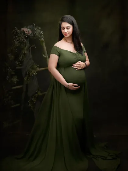 A full-length portrait in an olive green gown, set against a dark, moody backdrop with subtle floral elements. The lighting is carefully shaped to create a classic, painterly effect.