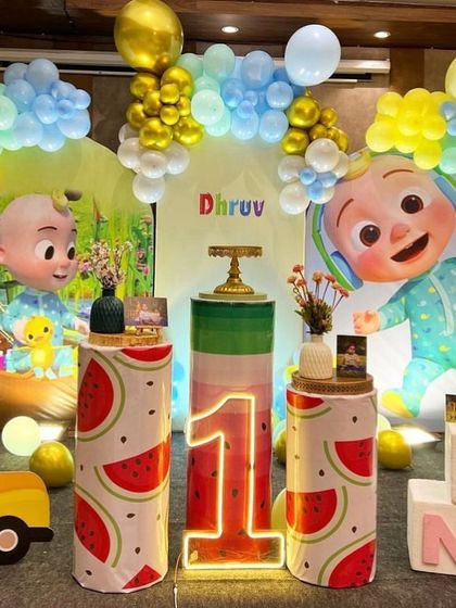 This Cocomelon setup for a first birthday features themed pedestals, a light-up number '1', and a yellow school bus prop, all framed by a blue and yellow balloon arch.