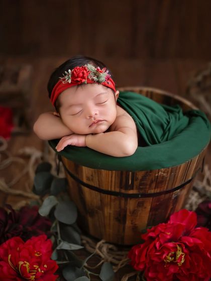 Rustic dreams and rosy cheeks. This baby girl is posed in a wooden bucket, wrapped in deep green and surrounded by vibrant red flowers.