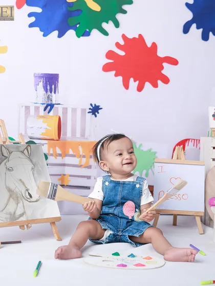 Holding two big paintbrushes, this little girl is ready to create. The vibrant paint splatters on the wall add a dynamic and fun element to the artist-themed photoshoot.