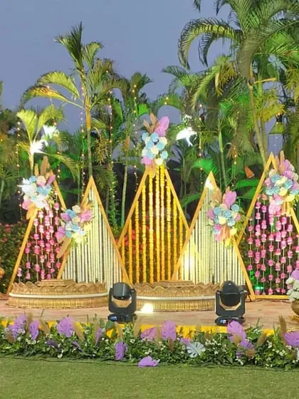 A grand outdoor Haldi stage setup. The triangular bamboo structures are decorated with floral arrangements and hanging strings, creating a beautiful backdrop against the natural greenery.