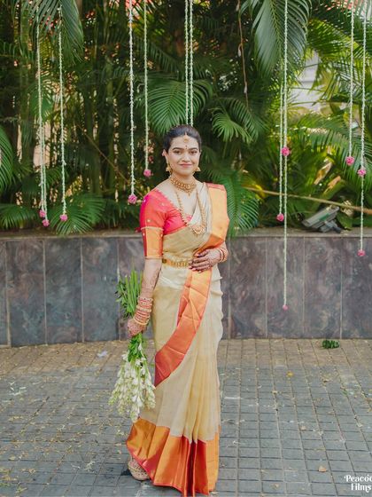A full-length portrait of the South Indian bride in her elegant off-white and orange saree, holding a bouquet of tuberoses.