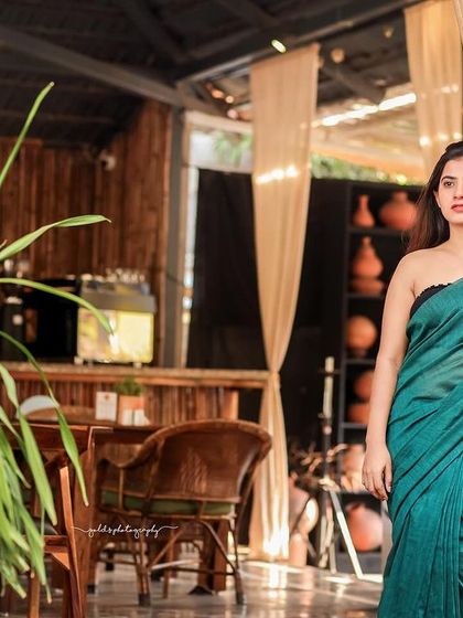 A modern take on saree styling. The model wears a teal saree paired with a contemporary black top, shot in a cafe with earthy tones. This shows how traditional wear can be styled for a modern, chic look.