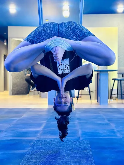 An inverted prayer pose, requiring balance, core strength, and concentration. The blue lighting of the studio creates a focused, serene atmosphere.