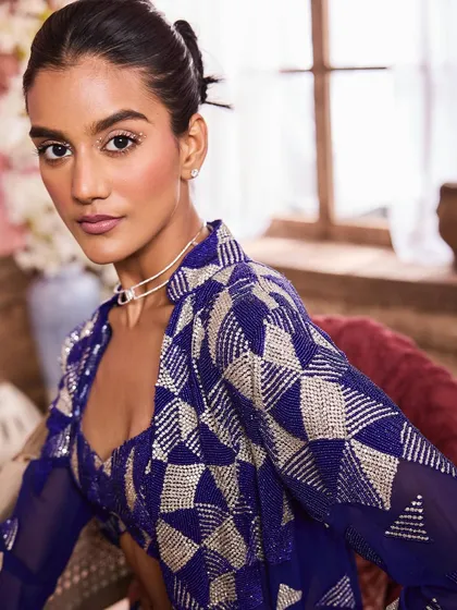 A close-up portrait showing a geometric print jacket. The makeup features a subtle shimmer on the eyelids and a clean, radiant skin finish.