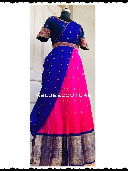 A full-length view of a classic half saree (langa voni) in a striking color combination. The ensemble features a pink skirt with a gold border, a royal blue dupatta, and a matching embroidered blouse.