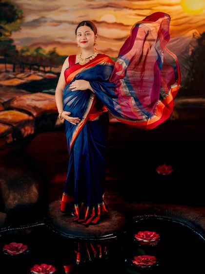 A full-length portrait of a mom-to-be in a blue and red saree, standing by a painted water scene. The flowing pallu adds a touch of dynamic energy.