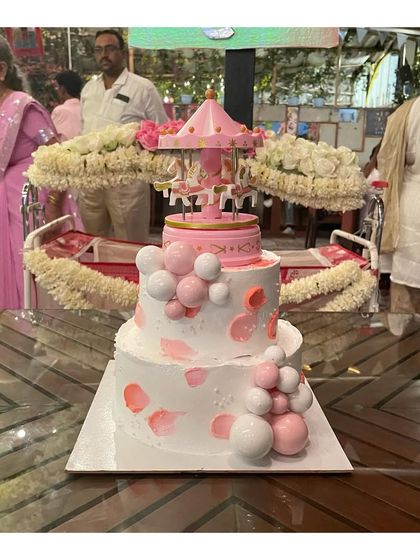 A lovely 2-tier cake for a baby's naming ceremony. The design features a rotating carousel on top and is decorated with pink and white chocolate spheres.