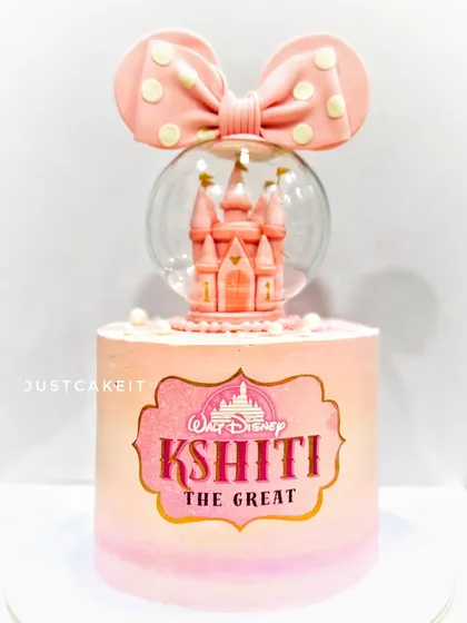 A magical Disney princess cake featuring a castle inside a snow globe. The pink ombre finish and a large polka-dot bow make it a dreamy centerpiece for a princess-themed party.