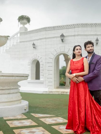 A grand, statuesque pose in front of a European-style fountain and building set. The woman's red dress adds a powerful splash of color.