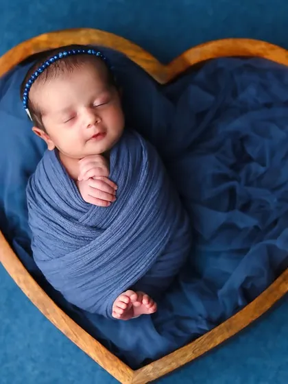 A beautiful composition featuring a newborn wrapped in deep blue fabric, nestled inside a wooden heart-shaped bowl. The rich color contrast creates a striking and lovely portrait.
