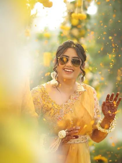 The bride, wearing cool sunglasses, laughs as she is showered with flower petals during her Haldi.