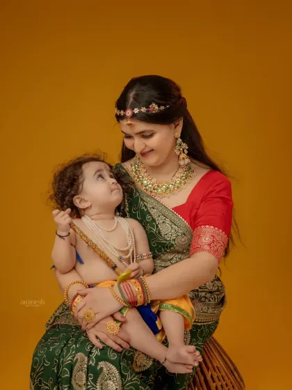 A tender moment captured between mother and child against a simple yellow backdrop. The baby, dressed as Krishna, looks up at his mother, creating an intimate and loving portrait.