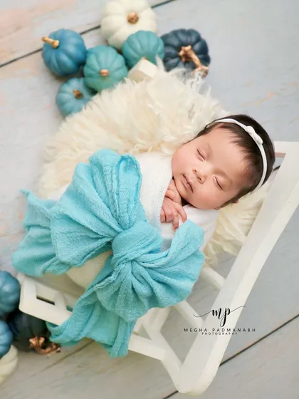 A baby rests on a miniature white bench, wrapped in a large aqua-blue bow. The blue and white pumpkins add a unique, seasonal touch.