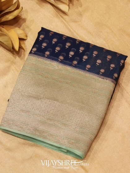 A dark blue Kora silk saree with a large silver zari border and delicate floral motifs. Kora silk is known for its texture and strength.