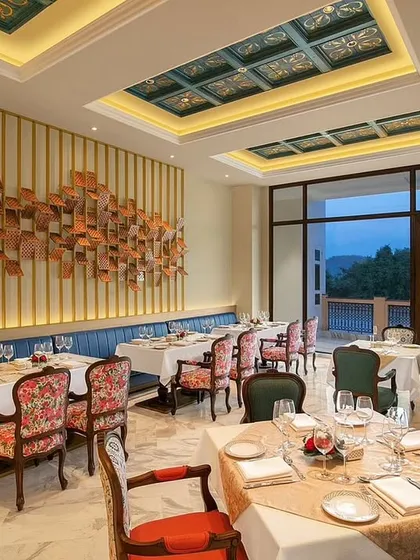 A dining area at IHCL Pilibhit House featuring a custom wall installation and floral upholstered chairs. The design balances a sophisticated blue banquette with vibrant, patterned seating to create a dynamic and visually rich dining space.