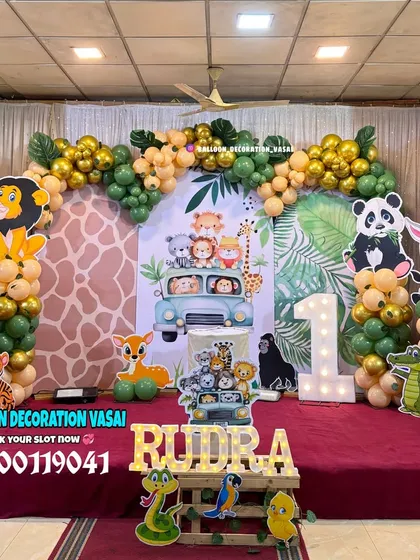 A full-stage jungle safari theme for a first birthday. This decoration includes a large backdrop, multiple animal cutouts, and a balloon arch in green and gold.
