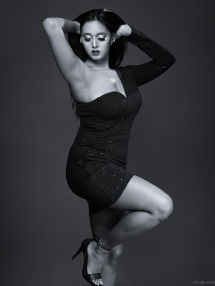 A dramatic monochrome portrait of a model in a one-shoulder dress. The lighting is sculpted to emphasize her pose and the texture of the fabric.