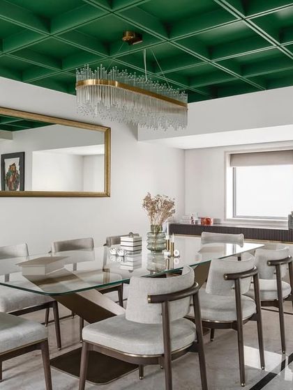 The dining room is a statement in itself. Crisp white chairs encircle a sleek glass-top table, all set beneath a striking green coffered ceiling with a dazzling chandelier that adds an aura of elegance.