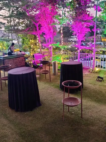 A practical setup for a cocktail party on a lawn, featuring standing cocktail tables with black covers and a fully equipped bar counter in the background, all under festive lighting.