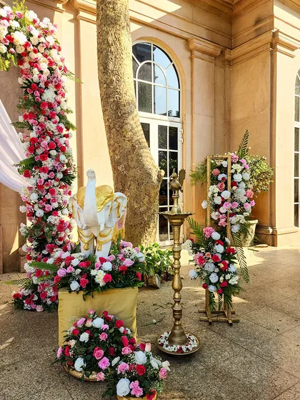 An outdoor decor setup for a traditional ceremony, featuring a floral arch of pink and white roses. The scene is accented with a classic brass lamp (diya) and a small elephant statue, blending sacred and celebratory elements.