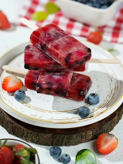 A stack of mixed berry popsicles on a rustic wooden platter, styled with fresh blueberries and strawberries for a farm-fresh feel.