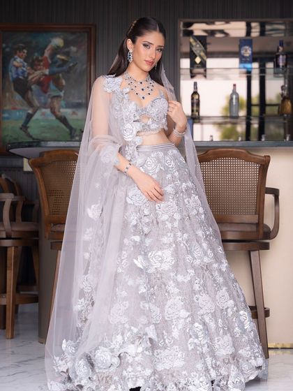 A full-length shot of a bride in a breathtaking silver-grey lehenga. The makeup is a subtle glam, designed to complement the cool tones of the outfit and the sparkle of the sapphire jewelry.