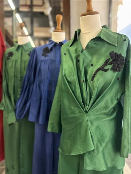 A collection of draped shirts in jewel tones of green and blue, featuring black floral appliqués and unique silhouettes.