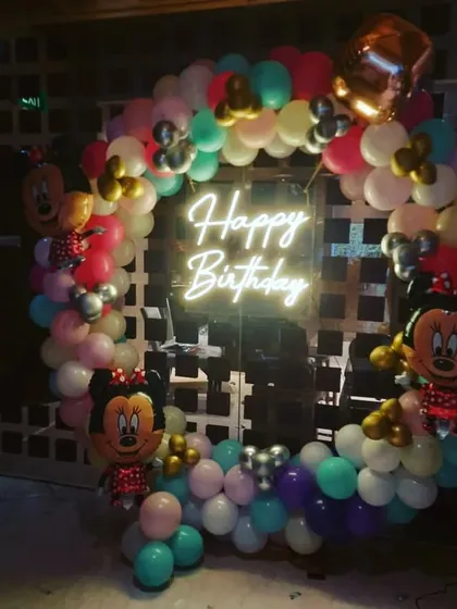 A classic Minnie Mouse themed birthday party. A circular balloon arch in pastel colors is decorated with Minnie Mouse foil balloons and a central 'Happy Birthday' neon sign.