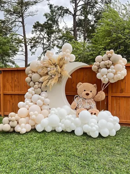 Welcoming a new baby is a joyous occasion. This outdoor 'Oh Baby' setup features a gentle, neutral color palette with a crescent moon backdrop, a cascade of balloons, and a cute teddy bear holding his own balloons.